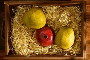 Two ripe pears and juicy pomegranate in a box with shavings on a wooden table, close-up, top view photo