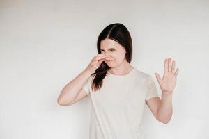 Woman dressed in casual white t shirt grimacing with disgust, holding breath, pinching nose with fingers to avoid bad smell and showing stop gesture, demonstrating repulsion to stink photo