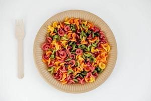 Multicolored pasta in a disposable paper plate with a wooden fork on a white background. Top view. Copy, empty space for text photo
