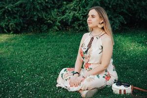 Beautiful blonde woman dressed in flower dress sitting on the green grass lawn. Copy, empty space for text photo