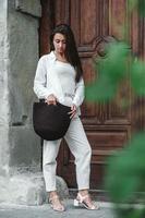 mujer vestida con ropa blanca, sosteniendo una bolsa de punto en el fondo de viejas puertas de madera. copiar, espacio vacío para texto
