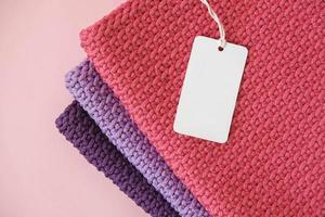 Stack of knitted material from threads of pink, red, purple colors with blank Price Tag on a pink background. Top view. Copy, empty space for text photo