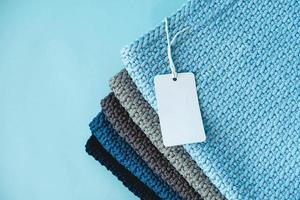 Stack of knitted material from threads of dark blue, light blue, gray colors with blank Price Tag on a blue background. Top view. Copy, empty space for text photo