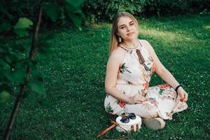 Beautiful blonde woman dressed in flower dress sitting on the green grass lawn. Copy, empty space for text photo
