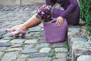 Woman wearing purple polka dot dress, holding knitted bag with flowers, posing in street of city. Copy, empty space for text photo