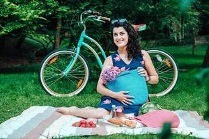 Woman dressed in a dress and with sunglasses holding a bag with flowers sitting on a plaid in park on background of bike photo