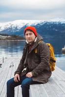 Man with a yellow backpack wearing a red hat sitting on wooden pier on the background of mountain and lake photo