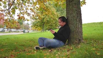 mujer asiática escribiendo y deslizando la pantalla en el teléfono inteligente en el parque en otoño, mujer sentada bajo el árbol y usando camisa negra de manga larga, hoja amarilla en el árbol, suecia