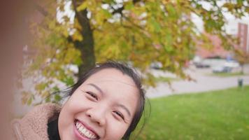 Close up of Happy Asian woman taking video call on smartphone at the park in Autumn, woman wearing light brown coat, yellow leaf on the tree, Beautiful day in Autumn season, River background in Sweden