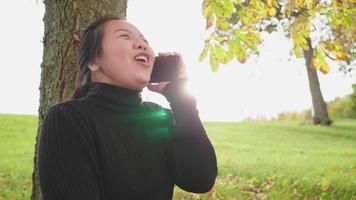 mujer asiática feliz sentada y haciendo una llamada en un teléfono inteligente bajo un árbol en el parque en otoño, usando una camisa negra de manga larga riéndose, hermoso día en la temporada de otoño con luz solar, suecia