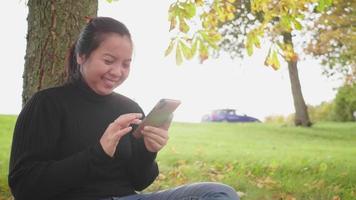 Aziatische vrouw zit en schuift het scherm op smartphone onder de boom in het park in de herfst, draagt een zwart shirt met lange mouwen en lacht, mooie dag met zonlicht, zweden, auto's op straat video