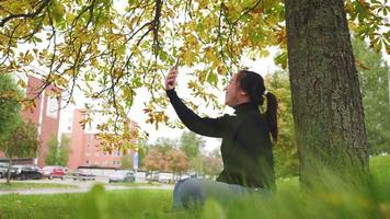 Asian woman sitting and taking a video call on smartphone under tree at the park in Autumn, woman wearing black long sleeved shirt, yellow leaf on the tree, Beautiful day in Autumn season, Sweden