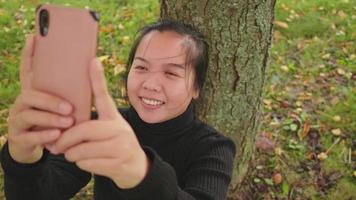 Top view of happy Asian woman standing and trying to take a picture of tree on smartphone, using smartphone taking a photo, yellow leaf on the ground, Beautiful day in Autumn season, sweden video