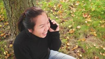 vista superior feliz mulher asiática sentada e tendo chamada no smartphone debaixo da árvore no parque no outono, vestindo camisa preta de manga comprida rindo, lindo dia na temporada de outono, suécia