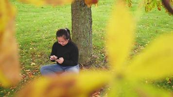 glad asiatisk kvinna som sitter och skjuter skärmen på smartphone under trädet i parken på hösten, kvinna klädd i svart långärmad skjorta, gult blad på trädet, sverige, tittar genom trädet