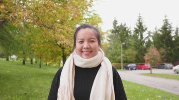 Front view of Asian woman standing and wearing black long-sleeved shirt and scarf at the park, woman looking at camera and smiling, yellow leaf on the tree, Beautiful day in Autumn season, Sweden video