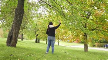 mujer asiática con camisa negra de manga larga y bufanda, tratando de tomar una foto de un árbol en el parque en otoño, usando un teléfono inteligente tomando una foto, hermoso día en la temporada de otoño, suecia video