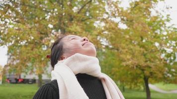 cerca de una mujer asiática feliz de pie y respirando profundamente en el parque en otoño, tomando un poco de aire fresco y sonriendo, hoja amarilla en el fondo del árbol, usando bufanda, suecia
