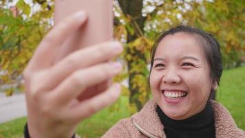 Close up of Happy Asian woman taking video call on smartphone at the park in Autumn, woman wearing light brown coat, yellow leaf on the tree, Beautiful day in Autumn season, Sweden
