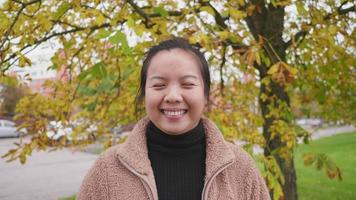 Front view of Asian woman standing and wearing light brown coat at the park in Autumn, woman looking at camera and smiling, yellow leaf on the tree, Beautiful day in Autumn season, Sweden video