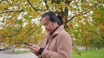 mujer asiática escribiendo y deslizando la pantalla en el teléfono inteligente en el parque en otoño, mujer de pie y usando un abrigo marrón claro, hoja amarilla en el árbol, hermoso día en la temporada de otoño, suecia video