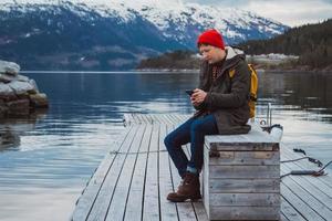 Man looking into the smartphone with a yellow backpack wearing a red hat sitting on wooden pier on background of mountain and lake