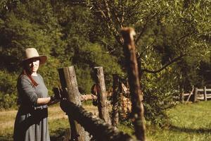 Woman wearing sundress and a straw hat standing near a wooden fence on a background of forest and trees at the sunset photo