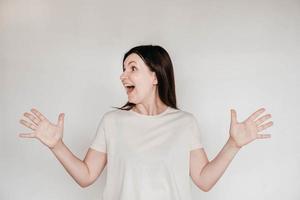 Emotional brunette woman with surprised face keeps holds hands to the side, dressed in casual white t shirt, poses indoor on a white background. Copy, empty space for text photo