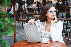mujer con gafas de sol y ropa blanca, sosteniendo una bolsa de punto sentada en la mesa de un café de la calle