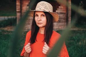 Portrait of a beautiful woman in a straw hat dressed in red clothes stands on the background of a wooden house photo