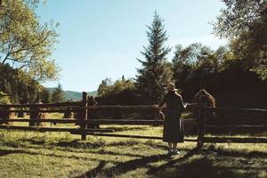 mujer con vestido de verano y sombrero de paja parada cerca de una valla de madera sobre un fondo de bosque, árboles, montañas y montones de heno al atardecer foto