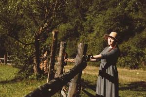 Woman wearing sundress and a straw hat standing near a wooden fence on a background of forest and trees at the sunset, with eyes closed photo