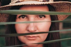 Portrait of a beautiful woman in a straw hat looking into the camera through the reed leaves photo