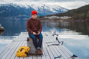 Man looking into the smartphone with a yellow backpack wearing a red hat sitting on wooden pier on the background of mountain and lake photo