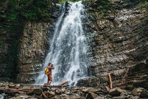 Traveler man with a yellow backpack standing on background of a waterfall makes a photo landscape
