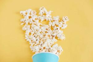 Popcorn in a blue paper cup scattered on a yellow background. Minimalist concept. Top view. Copy, empty space for text photo