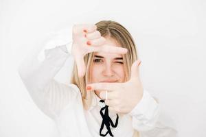Young beautiful blonde woman wearing stylish white shirt making frame with her hands on a white wall background. Copy, empty space for text photo
