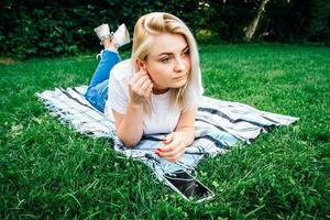 hermosa mujer rubia con auriculares y con un smartphone en las manos escuchando música tirada en el césped al aire libre en el parque. lugar para texto o publicidad foto