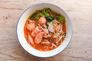 Red soup noodle pork with red roasted pork and bean sprout vegetable in soup bowl on wooden table, top view Thai and China Asian food photo