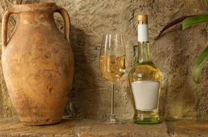 old clay jug and a bottle of white wine with a wineglass stand on the background of a clay wall photo