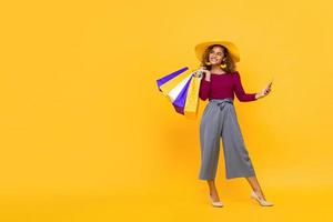 retrato completo de una mujer afroamericana sonriente sosteniendo bolsas de compras y un teléfono móvil mientras mira de lado para copiar el espacio en el fondo amarillo foto