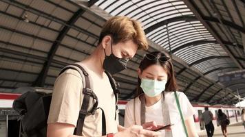 Couple Asian tourists with face masks search information, find travel locations by mobile phone map at a train station in Thailand, passenger trip lifestyle, casual transportation, journey vacation.
