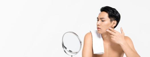 Shirtless young handsome Asian man looking at his face in the mirror  isolated on white banner background for skin care and beauty concepts