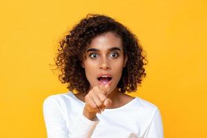 primer plano retrato de una atractiva joven afroamericana que parece sorprendida mientras señala con el dedo al frente en un estudio aislado de fondo amarillo foto