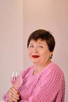 alegre mujer madura en suéter rosa bebiendo vino de rosas y sonriendo. autosuficiencia, vacaciones, concepto de celebración