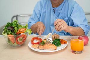 Asian senior or elderly old lady woman patient eating Salmon steak breakfast with vegetable healthy food while sitting and hungry on bed in hospital. photo