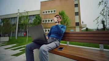 joven que trabaja en la computadora portátil al aire libre y bebe café. video