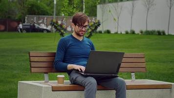 Young man in city in park typing laptop chat working remotely e-learning online. video