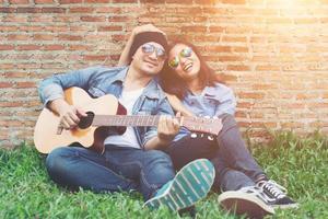 hombre hipster tocando la guitarra para su novia al aire libre contra la pared de ladrillo, disfrutando juntos. foto