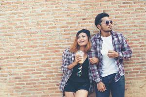 Portrait of young hipster couple posing with her boyfriend holding a cup of coffee enjoying holiday together against brick wall background. photo